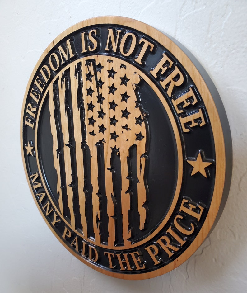 K&ouml;nnte beinhalten: Ein rundes Holzschild mit schwarzem Hintergrund und einem verwitterten amerikanischen Flaggenmuster. Der Text "Freedom is not free, many paid the price" ist in das Holz geschnitzt.