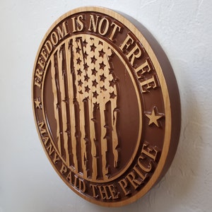 K&ouml;nnte beinhalten: Ein rundes Holzschild mit braunem Hintergrund und einer geschnitzten amerikanischen Flagge in der Mitte. Das Schild lautet "Freiheit ist nicht umsonst, viele haben den Preis bezahlt".
