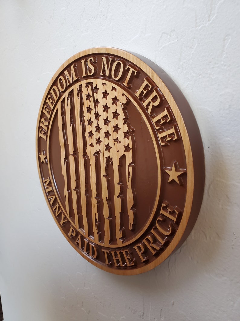 K&ouml;nnte beinhalten: Ein rundes Holzschild mit braunem Hintergrund und einer geschnitzten amerikanischen Flagge in der Mitte. Das Schild lautet "Freiheit ist nicht umsonst, viele haben den Preis bezahlt".