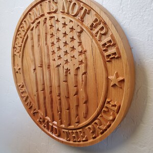 K&ouml;nnte beinhalten: Ein rundes Holzschild mit dem Text "Freedom is not free, many paid the price", der in das Holz geschnitzt ist. Das Schild zeigt au&szlig;erdem eine geschnitzte amerikanische Flagge mit Sternen und Streifen.