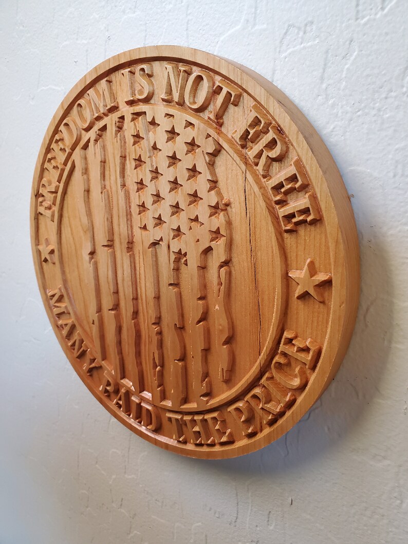 K&ouml;nnte beinhalten: Ein rundes Holzschild mit dem Text "Freedom is not free, many paid the price", der in das Holz geschnitzt ist. Das Schild zeigt au&szlig;erdem eine geschnitzte amerikanische Flagge mit Sternen und Streifen.