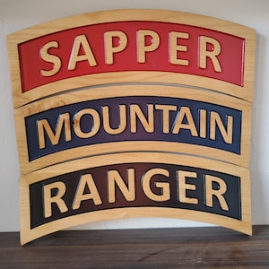 May include: Three wooden plaques with curved edges, each with a different military insignia. The top plaque is red with the word "SAPPER" in yellow lettering. The middle plaque is blue with the word "MOUNTAIN" in yellow lettering. The bottom plaque is black with the word "RANGER" in yellow lettering.