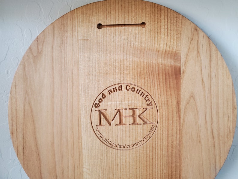 K&ouml;nnte beinhalten: Ein rundes Holzschild mit einem kreisf&ouml;rmigen Logo, auf dem "God and Country" steht, mit den Initialen "MHK" im Kreis. Das Logo enth&auml;lt auch die Website "www.godandcountryetsy.com".