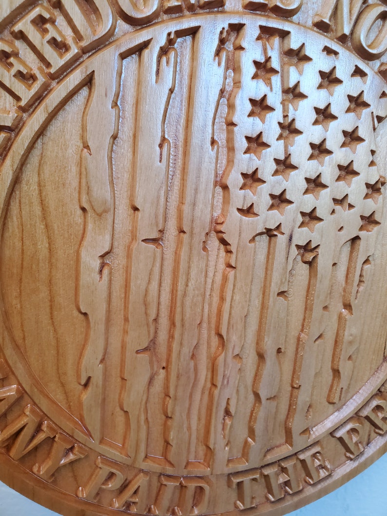 K&ouml;nnte beinhalten: Ein rundes Holzschild mit einem geschnitzten amerikanischen Flaggenmuster. Das Schild tr&auml;gt die Aufschrift "ICH HABE DEN PREIS F&Uuml;R MEINE FREIHEIT GEZAHLT".