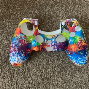 May include: A colourful controller skin with a vibrant splatter paint design. The skin features a white interior and a pattern of red, blue, yellow, and green paint splatters. This accessory is designed for gaming.