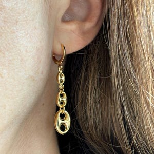 May include: Gold-colored dangle earrings with a coffee bean design. The earrings are made of a shiny, metallic material and feature a small hoop that attaches to the ear. The earrings have three connected coffee bean-shaped charms.