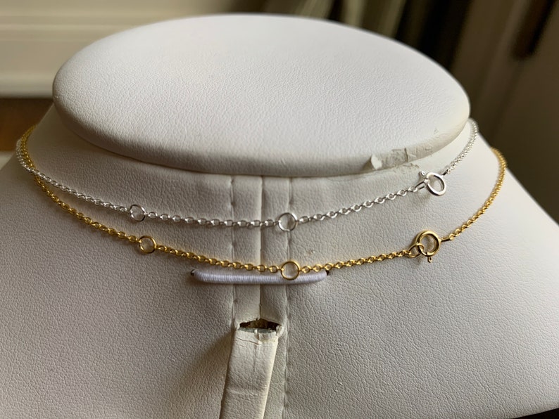 May include: Two necklaces on a white mannequin bust. The top necklace is silver and the bottom necklace is gold.