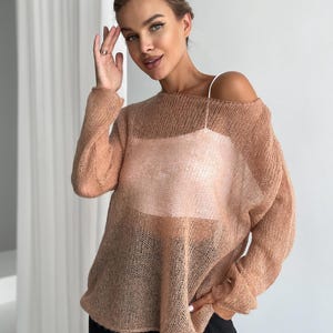 May include: A peach-coloured, open-knit sweater with long sleeves. The sweater is worn over a white camisole and paired with black trousers. The sweater has a loose, relaxed fit.