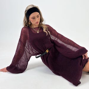 Könnte beinhalten: Burgunderfarbenes Kleid mit langen Ärmeln und offenem Strickmuster. Das Kleid wird mit einem schwarzen Gürtel mit goldfarbener Schnalle in der Taille zusammengehalten. Das Model trägt ein schwarzes Stirnband und Goldschmuck.