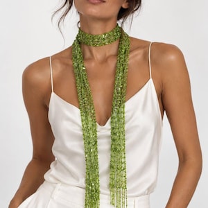 May include: A long, lime green scarf featuring a delicate, open-knit mesh construction adorned with numerous small, shimmering sequins. The scarf is wrapped around the neck and hangs down in two long, slender, fringed panels over a white camisole.