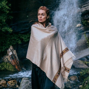 May include: A woman wearing a beige shawl with a colorful patterned border stands in front of a waterfall. The shawl is draped over her shoulders and she is looking off to the side.