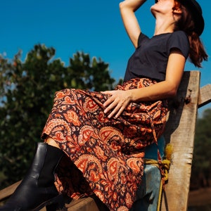 May include: A woman wearing a black t-shirt and a long, flowing skirt with a paisley print sits on a wooden structure. She is wearing a black hat and has her hand on her hat.