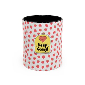 May include: A ceramic mug with a black interior and a white exterior featuring a repeating pattern of small red strawberries on a light grid background. A central yellow arch-shaped label displays a red heart with a face and the text Keep Going!