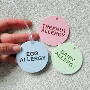 May include: Three round allergy alert tags with a clear string. The tags are light blue, pink, and green. The text on the tags reads "Egg Allergy", "Tree Nut Allergy", and "Dairy Allergy".