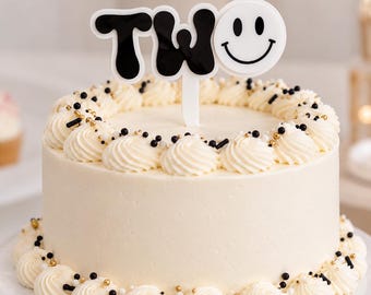 May include: A round cake with smooth white frosting, decorated with piped white frosting swirls and small black and gold sprinkles. A cake topper displays the word TWO in black letters, with the letter O replaced by a white smiley face icon.