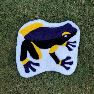May include: A white rug featuring a purple and black frog design. The frog has yellow accents on its body and feet.