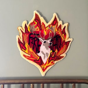 May include: A heart-shaped rug with a deer head in the center, surrounded by flames. The rug is made of yarn and has a textured, fluffy surface. The flames are in shades of red, orange, and yellow. The deer head is white with brown antlers. The text "FIRE HEART" is written in black letters on the rug.