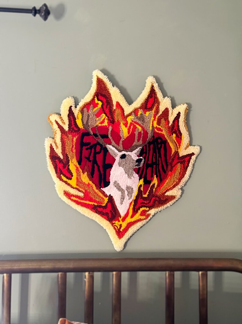 May include: A heart-shaped rug with a deer head in the center, surrounded by flames. The rug is made of yarn and has a textured, fluffy surface. The flames are in shades of red, orange, and yellow. The deer head is white with brown antlers. The text "FIRE HEART" is written in black letters on the rug.