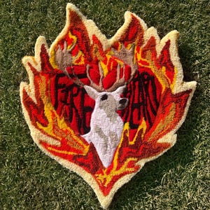 May include: A heart-shaped rug with a deer head in the center, surrounded by flames in shades of red, orange, and yellow. The rug is made of a fluffy, textured material.