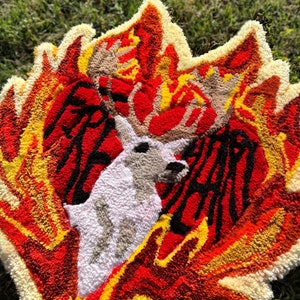 May include: A white deer with large antlers is surrounded by flames in a rug made of yarn. The flames are red, orange, and yellow. The rug is a unique and colorful piece of art.