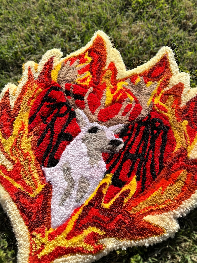 May include: A white deer with large antlers is surrounded by flames in a rug made of yarn. The flames are red, orange, and yellow. The rug is a unique and colorful piece of art.