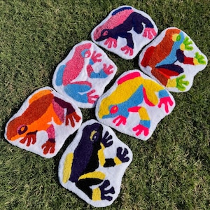 Puede incluir: Una colección de cinco tapices o alfombras textiles con mechones, cada uno con forma de rana. Las ranas presentan patrones vibrantes y multicolores que incluyen rosa, azul, naranja, amarillo, morado y negro, sobre un fondo blanco.