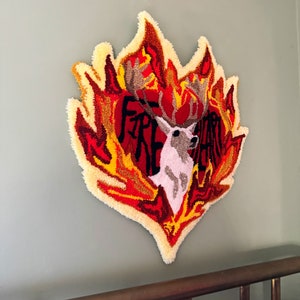 May include: A heart-shaped wall hanging with a deer in the center, surrounded by flames in shades of red, orange, and yellow. The word "FIRE" is visible in the heart.