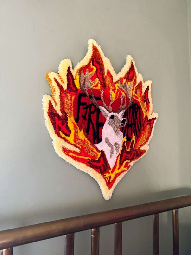May include: A heart-shaped wall hanging with a deer in the center, surrounded by flames in shades of red, orange, and yellow. The word "FIRE" is visible in the heart.