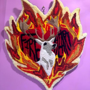 May include: A heart-shaped rug with a deer head in the center and the words "Fire Heart" written around it. The rug is made of yarn and has a flame-like design in red, yellow, and orange.
