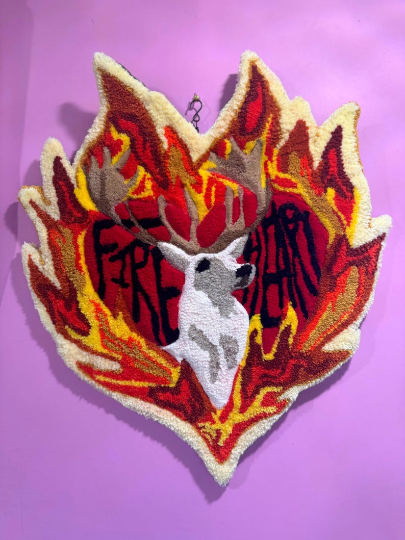May include: A heart-shaped rug with a deer head in the center and the words "Fire Heart" written around it. The rug is made of yarn and has a flame-like design in red, yellow, and orange.
