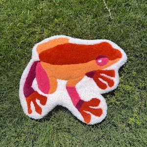 Puede incluir: Una alfombra blanca y esponjosa con forma de rana con detalles en naranja, rosa y morado. La rana está mirando hacia la derecha y tiene un ojo grande y redondo.