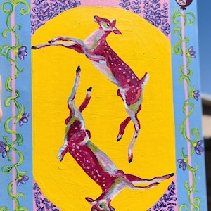 May include: An acrylic painting on canvas featuring two leaping deer with red and white spotted coats against a yellow circle. The background has a blue and pink border with floral and pomegranate details.