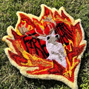 May include: A red, yellow, and white rug with a deer head in the center. The deer head is surrounded by flames. The rug is made of yarn and has a fluffy texture.