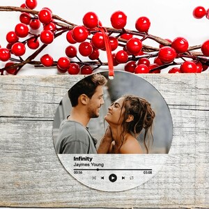 Peut inclure: Une boule de No&euml;l ronde et argent&eacute;e avec une photo d'un couple se regardant. La boule a un design de lecteur de musique avec le titre de la chanson "Infinity" de Jaymes Young.