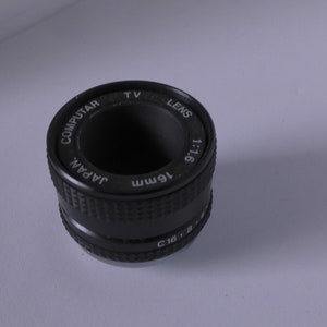 Può includere: Un obiettivo nero da 16 mm con la scritta "Computar TV Lens 1:1.6 16mm Japan" stampata attorno al barilotto.