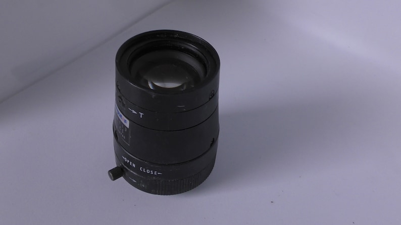 K&ouml;nnte beinhalten: Ein schwarzes CCTV-Objektiv mit einem Fokusring und einem Zoom-Einstellknopf. Das Objektiv hat einen schwarzen Kunststoffk&ouml;rper und eine klare Glaslinse.