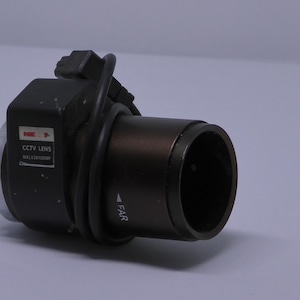 May include: A black CCTV lens with a silver ring and a black cable. The lens has a label that reads "CCTV LENS" and "MULTI-COATED". The lens has a "FAR" label on the side.