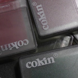 Op de afbeelding: Close-up van vier grijze plastic filterhouders met het woord "Cokin" erop gedrukt. De houders zijn op elkaar gestapeld.