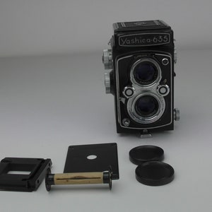 May include: A black and silver Yashica 635 camera with two lenses, a film roll, a film back, and two lens caps.