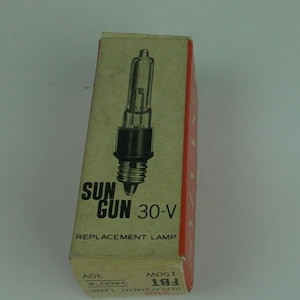 Può includere: Una scatola di cartone con un'etichetta rossa e bianca. L'etichetta dice "Sun Gun 30-V Replacement Lamp". La scatola contiene una lampadina di vetro trasparente con una base nera.