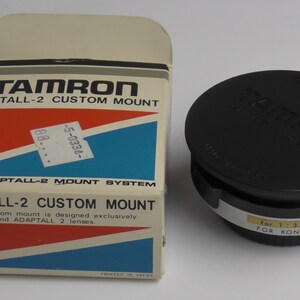 May include: A black Tamron Adaptall-2 custom mount adapter for Konica cameras. The adapter is in a box with the text "Tamron Adaptall-2 Custom Mount" and "For Adaptall-2 Mount System".
