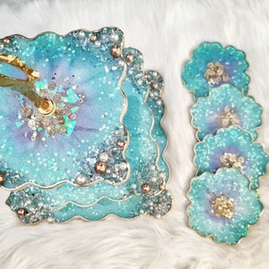 Op de afbeelding: Een drielaags taartstandaard met een gouden handvat. Elke laag is gemaakt van helder hars met een blauwe en paarse swirl en glitter. De standaard is versierd met parels en gouden accenten. Er zijn vier bijpassende onderzetters met hetzelfde ontwerp.