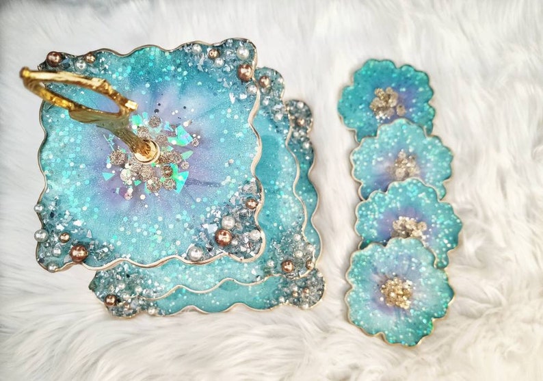 Op de afbeelding: Een drielaags taartstandaard met een gouden handvat. Elke laag is gemaakt van helder hars met een blauwe en paarse swirl en glitter. De standaard is versierd met parels en gouden accenten. Er zijn vier bijpassende onderzetters met hetzelfde ontwerp.