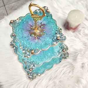 Op de afbeelding: Een drielaags taartstandaard met een gouden handvat. Elke laag is gemaakt van hars en heeft een blauw en paars swirl-ontwerp met glitter- en parel accenten.