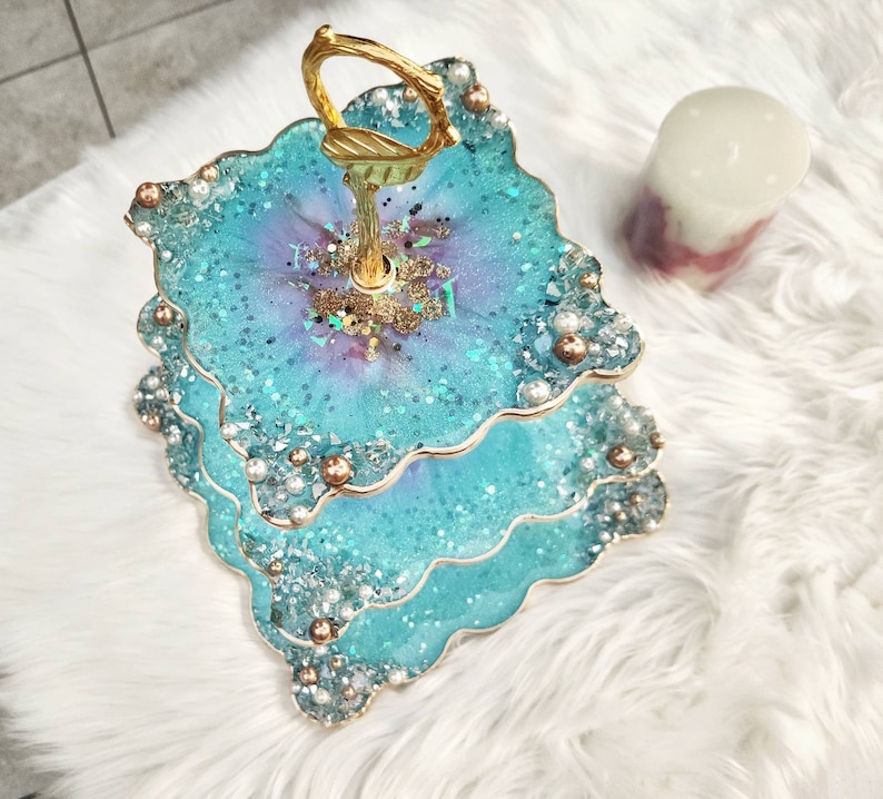 Op de afbeelding: Een drielaags taartstandaard met een gouden handvat. Elke laag is gemaakt van hars en heeft een blauw en paars swirl-ontwerp met glitter- en parel accenten.