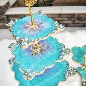 Op de afbeelding: Een drielaags taartstandaard gemaakt van turquoiseblauwe hars met een bladgoudontwerp en parel accenten. De standaard heeft een gouden metalen handvat en is perfect voor het presenteren van desserts of andere lekkernijen.