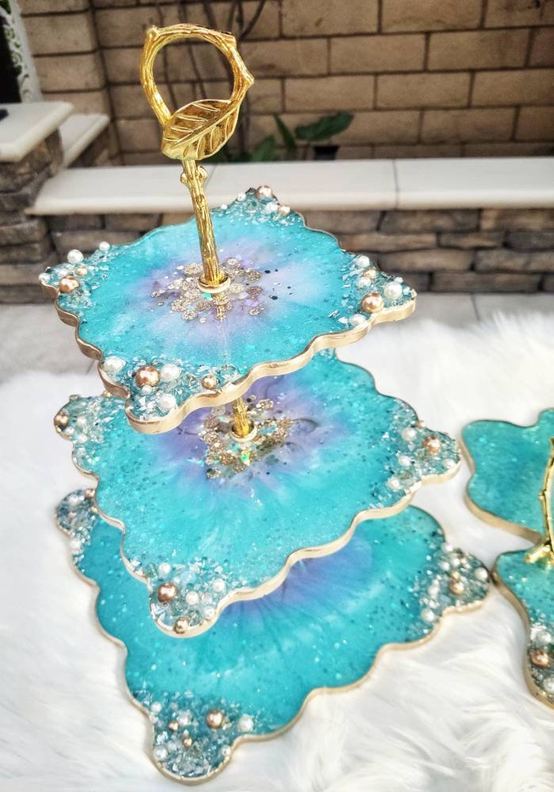 Op de afbeelding: Een drielaags taartstandaard gemaakt van turquoiseblauwe hars met een bladgoudontwerp en parel accenten. De standaard heeft een gouden metalen handvat en is perfect voor het presenteren van desserts of andere lekkernijen.