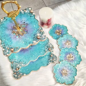 Op de afbeelding: Een tweelaagse taartstandaard met een gouden metalen frame en blauwe en paarse harsbladen versierd met glitter en parels. De standaard is omgeven door vijf kleinere blauwe en paarse harsonderzetters met glitter en parels.