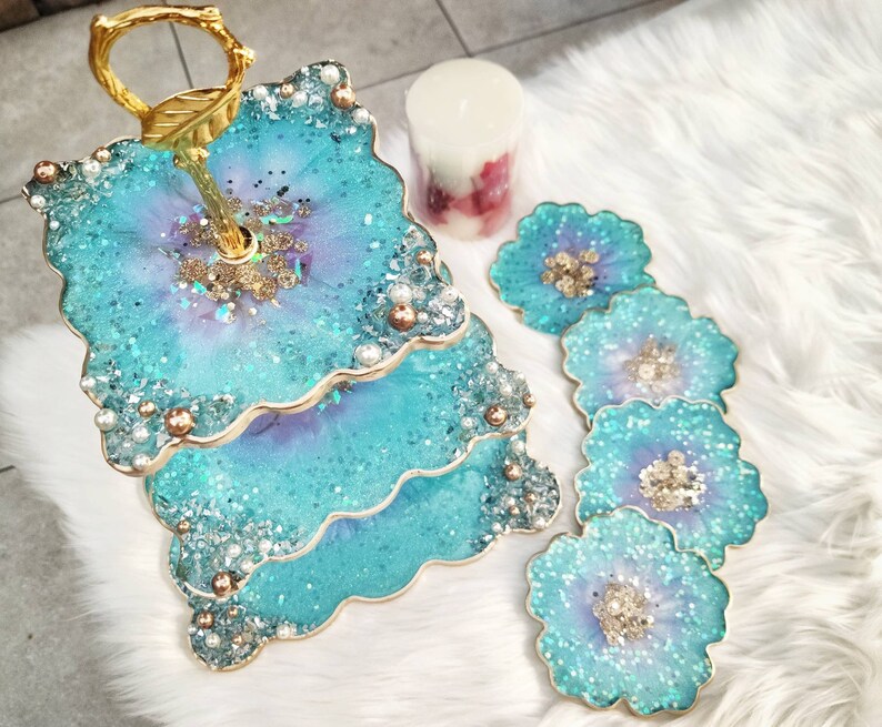 Op de afbeelding: Een tweelaagse taartstandaard met een gouden metalen frame en blauwe en paarse harsbladen versierd met glitter en parels. De standaard is omgeven door vijf kleinere blauwe en paarse harsonderzetters met glitter en parels.