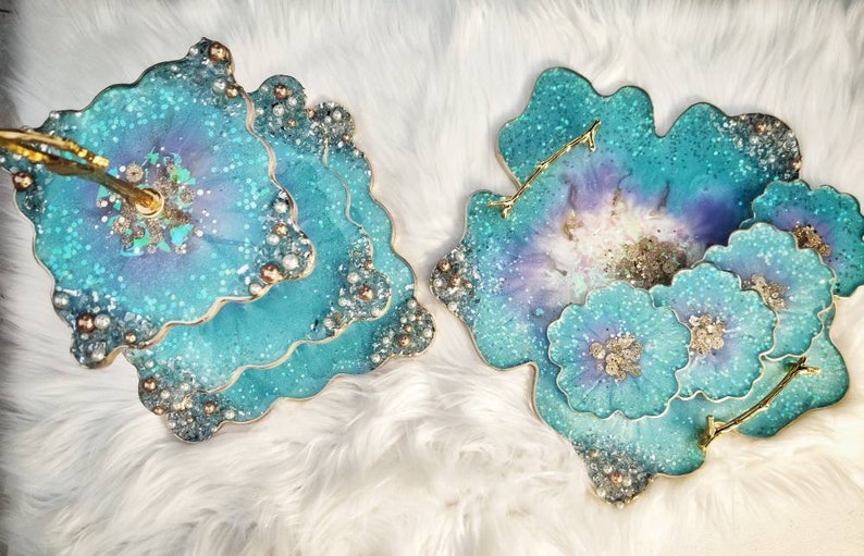 Op de afbeelding: Drie-laags turquoiseblauwe hars serveerplateau met een gouden standaard en parel accenten. Het plateau heeft een unieke, organische vorm en is perfect om desserts, hapjes of andere lekkernijen te presenteren.